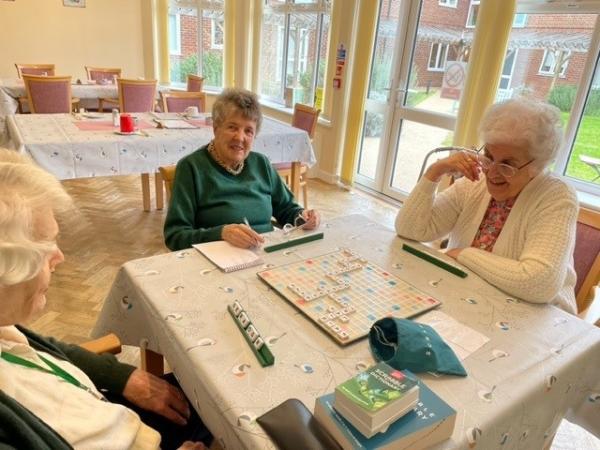 three smiling older people playing scrabble in a large, homely dining room.