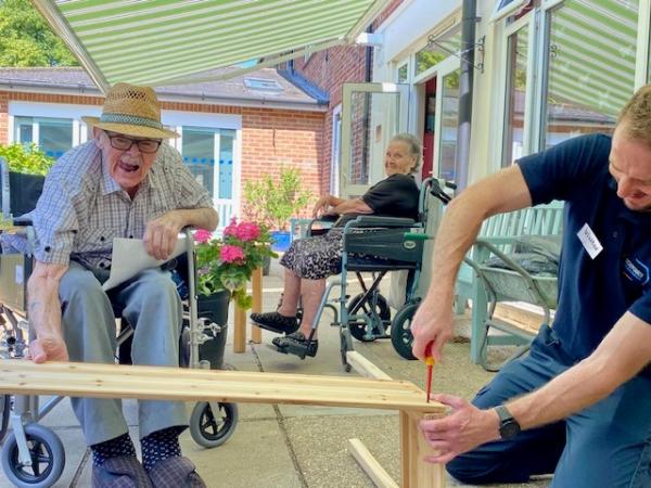 A resident and volunteer build a planter while another resident watches, smiling