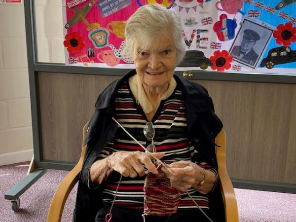 Smiling resident at Corton House knitting. In the background is a bright paintin