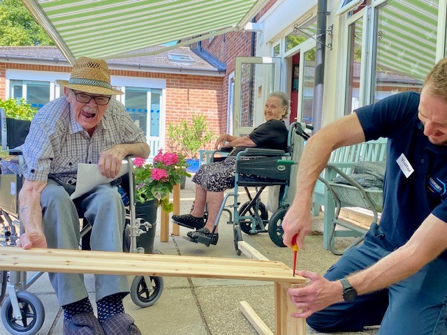 A resident and volunteer build a planter while another resident watches, smiling