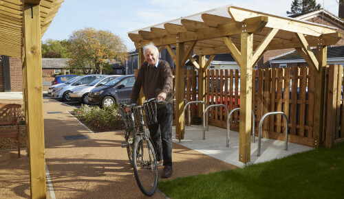 A tenant at Brakendon Close Independent Living Scheme in Norwich stands in the garden and car park area of the sheltered housing complex, smiling with his bike next to a purpose built bike shed.