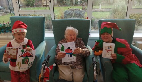 A resident in Norwich care home Corton House smiles with her penpals, local children Kieran and Emily, who are dressed in Christmas outfits. All three are holding festive artwork.